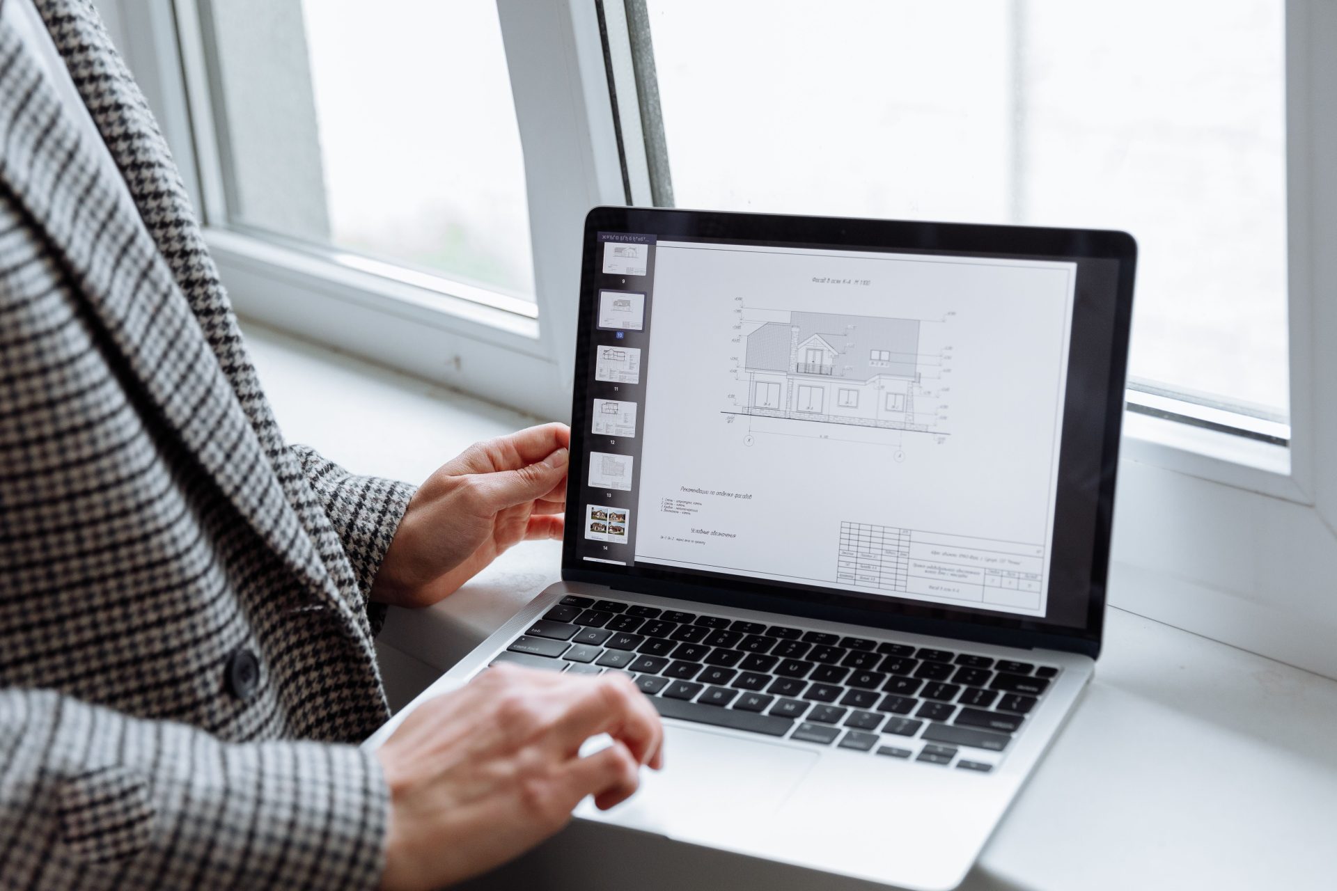 Person in plaid blazer reviewing architectural blueprints on laptop by a window.